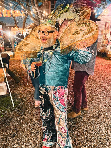 Person in elaborate costume with large hat and glasses, holding a drink, on a street.
