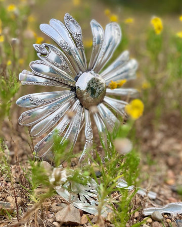 Decorative silver flower made from cutlery in a natural setting with greenery and small yellow flowers.