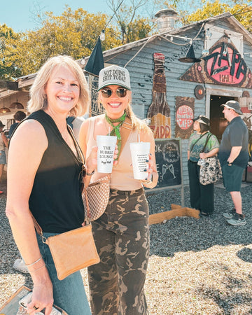 Two women standing outdoors with a rustic building and people in the background