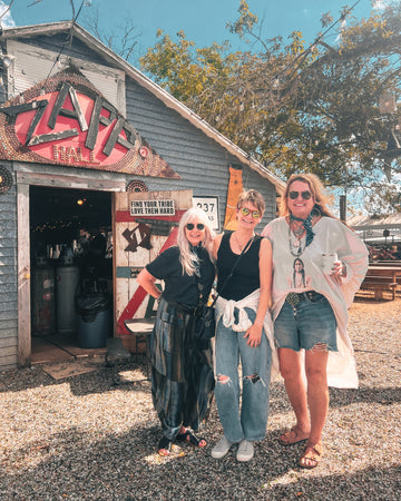 Three women standing in front of a rustic building with a sign that reads 'Zap'.