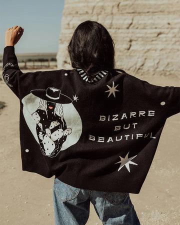 Person wearing a black jacket with a graphic design and text, standing in front of a stone wall.