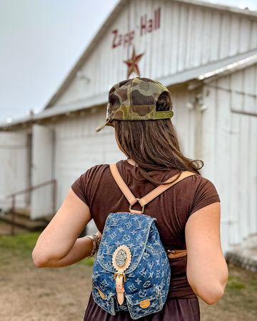 Person wearing a camouflage cap and Leatherandvodka denim backpack in front of a building with 'Zapp Hall' sign.