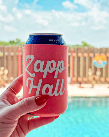 Hand holding a pink can cooler with 'Zapp Hall' branding by a pool