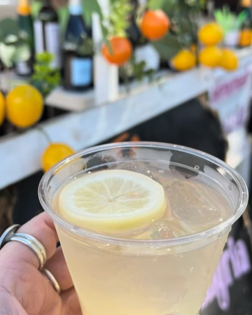 Clear plastic cup with lemon slice held by a hand, blurred background with bottles and fruits