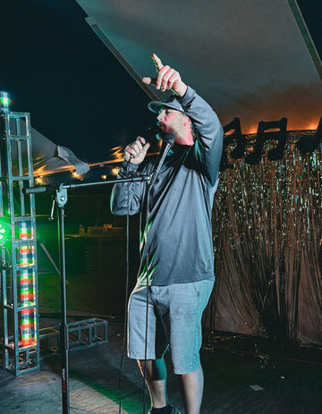 Person on stage with microphone, wearing a cap and casual clothing, against a dark background.
