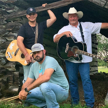 Three men with guitars outdoors, one holding drumsticks, against a rustic stone wall.