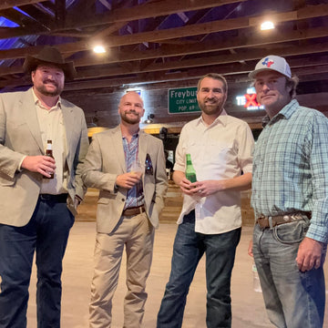 Four men standing together in a barn-like setting, holding drinks.