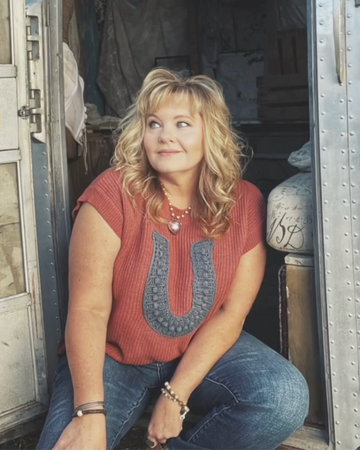 Woman sitting inside a vintage trailer wearing a red top with blue patterns and jeans.