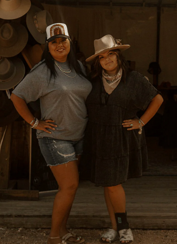 Two women posing together in front of a wall of hats.