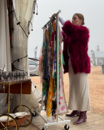 Woman in a fur coat standing next to a rack of colorful clothing on a wooden platform.