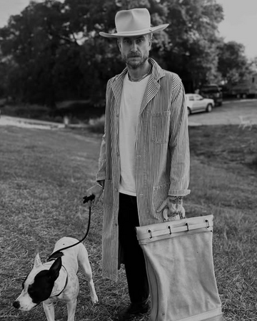 Man in a long coat and hat walking a dog in a field