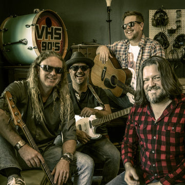 Four musicians posing with their instruments in a room with a VHS sign in the background