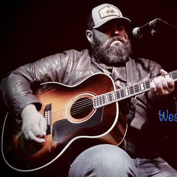 Man playing an acoustic guitar with a microphone, wearing a cap and leather jacket.